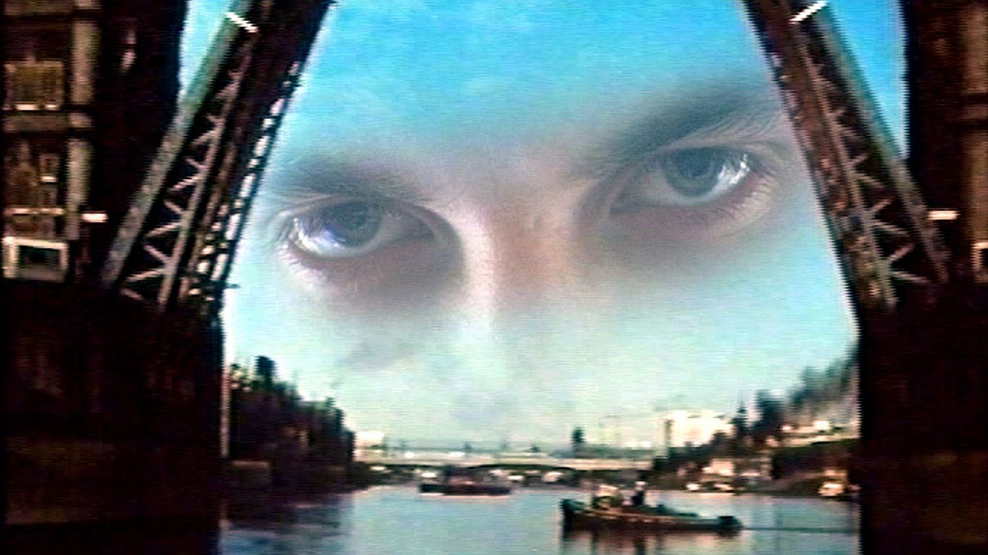 This film still shows two superimposed shots of London Tower Bridge, which has opened its flaps to allow larger ships to pass through. In the background, there is actually a smaller steamboat from which black smoke is coming out. In the upper part of the picture, there is in turn a pair of eyes that look intensively into the camera and are flanked by the open locks.