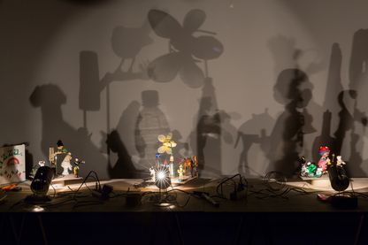Installationsaufnahme der Arbeiten "Schatten" des Künstlers Hans-Peter Feldmann. Dabei handelt es sich um eine Arbeit, bestehend aus verschiedenen kleineren Artefakten auf einem Tisch, die von mehreren kleinen Lichtquellen angeleuchtet werden und so einen Schatten auf die dahinter liegende Wand werfen. Hans-Peter Feldmann, Sammlung Goetz München