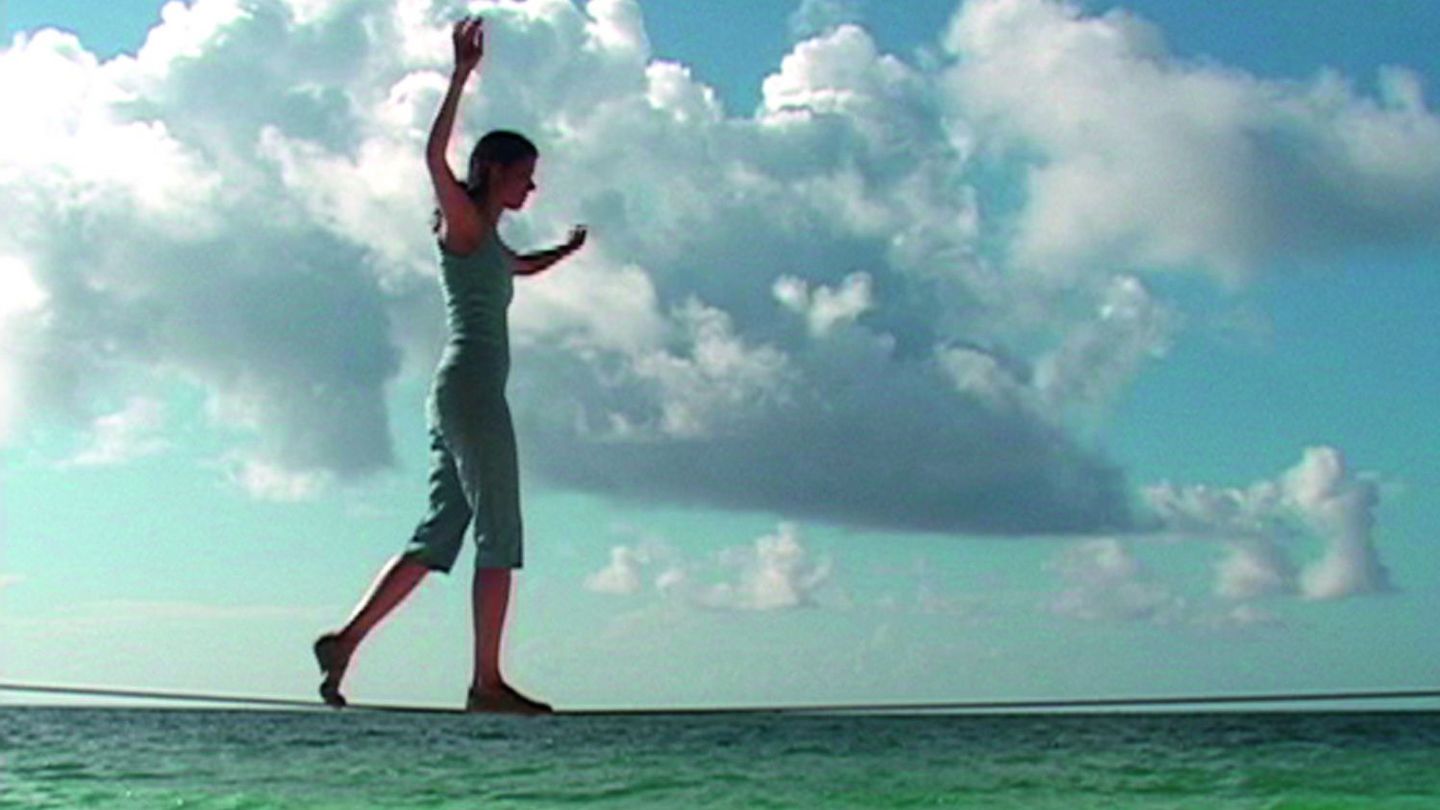 Dieses Video Still zeigt ein Frau in sportlicher Kleidung im Profil, die vor einem Horizont mit Meer und leicht bewölktem Himmel auf einem Seil genau auf der Horizontlinie balanciert. Janine Antoni, Sammlung Goetz München