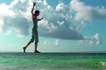 Dieses Video Still zeigt ein Frau in sportlicher Kleidung im Profil, die vor einem Horizont mit Meer und leicht bewölktem Himmel auf einem Seil genau auf der Horizontlinie balanciert. Janine Antoni, Sammlung Goetz München