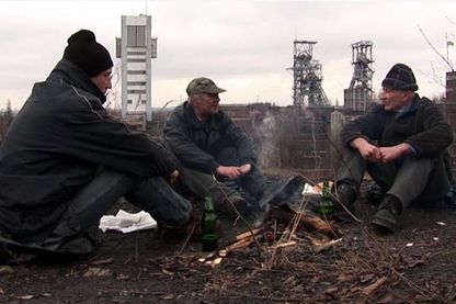 Das Video Still zeigt drei Männer unterschiedlichen Alters in Winterkleidung an einem grauen Tag um ein Feuer herum sitzend. Sie scheinen sich miteinander zu unterhalten. Im Hintergrund ist eine Art Industriegebiet zu sehen. Anna Molska, Sammlung Goetz München
