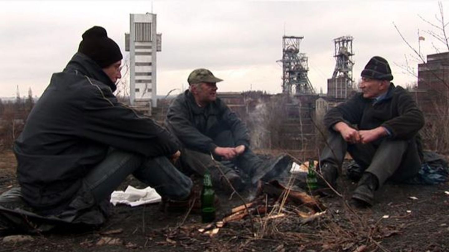 Das Video Still zeigt drei Männer unterschiedlichen Alters in Winterkleidung an einem grauen Tag um ein Feuer herum sitzend. Sie scheinen sich miteinander zu unterhalten. Im Hintergrund ist eine Art Industriegebiet zu sehen. Anna Molska, Sammlung Goetz München
