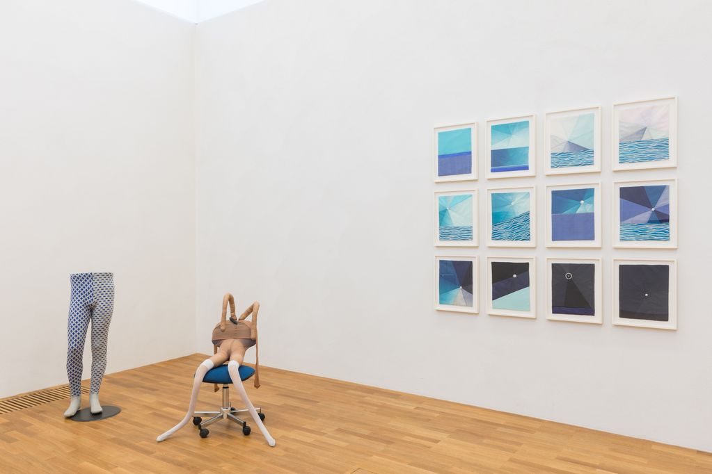 Exhibition view with blue-grey woollen tights on male mannequin legs, a human-like structure made of silk tights on a blue office chair, and twelve small-format blue fabric works in white frames on the wall. Rosemarie Trockel/Sarah Lucas/Louise Bourgeois, Sammlung Goetz Munich