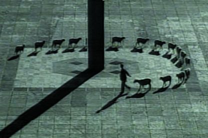 Auf diesem Filmstill in schwarz-weiß erkennt man einen Mann aus der Vogelperspektive, der auf einem steinernen Platz eine Schafherde um eine große Säule führt. Die Schafe sind dabei ordentlich hintereinander aufgereiht und werfen lange Schatten.