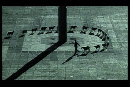 In this black and white film still, a man can be seen from a bird's eye view, leading a flock of sheep around a large column in a stone square. The sheep are neatly lined up one behind the other and are casting long shadows.