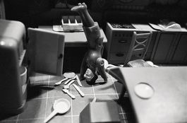Black-and-white photograph of a Playmobil doll in a dollhouse kitchen. The doll is doing a handstand, and plates and cutlery are lying on the kitchen floor next to her