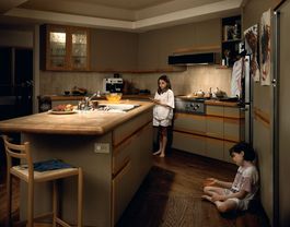 The picture shows a spacious kitchen with a centrally located kitchen island. On the right side of the frame, a girl is sitting cross-legged on the floor, leaning against a refrigerator. The way she moves her hand gives the impression that she is explaining or telling a story. Behind the kitchen island stands another girl, leaning against the stove. Her gaze is directed into the room, while her posture appears pensive or lost in thought.