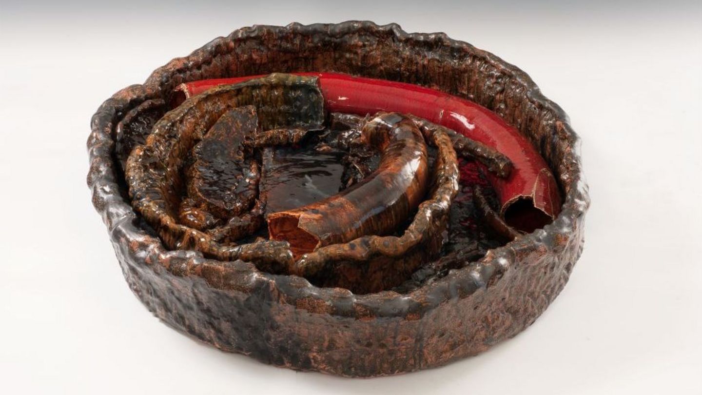 A large clay bowl is filled with sausage-shaped ceramic elements. The colouring shifts between brown and red. The surfaces display an irregular pattern, and the glaze is highly glossy.