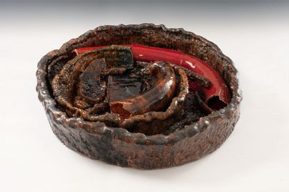 A large clay bowl is filled with sausage-shaped ceramic elements. The colouring shifts between brown and red. The surfaces display an irregular pattern, and the glaze is highly glossy.