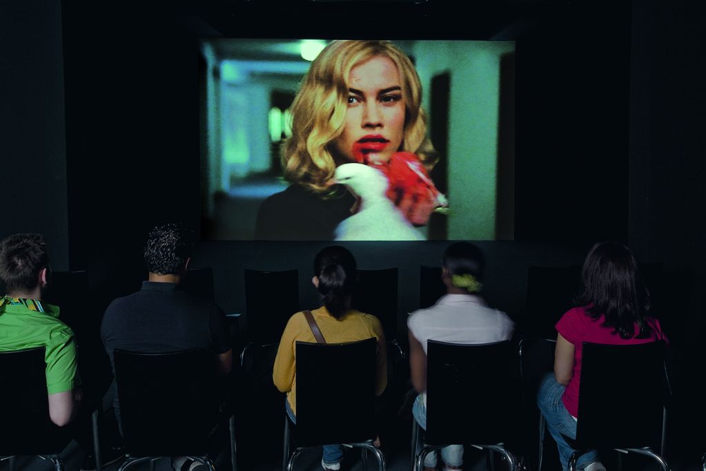 This installation view shows the screening of the video work "Looking for Alfred" by Johan Grimonprez. Five different people are seen sitting on black chairs with their backs to the viewer, the still image of the video shows a white woman with blonde hair and a bloodied mouth who appears to be eating the insides of a white pigeon. Johan Grimonprez, Sammlung Goetz Munich