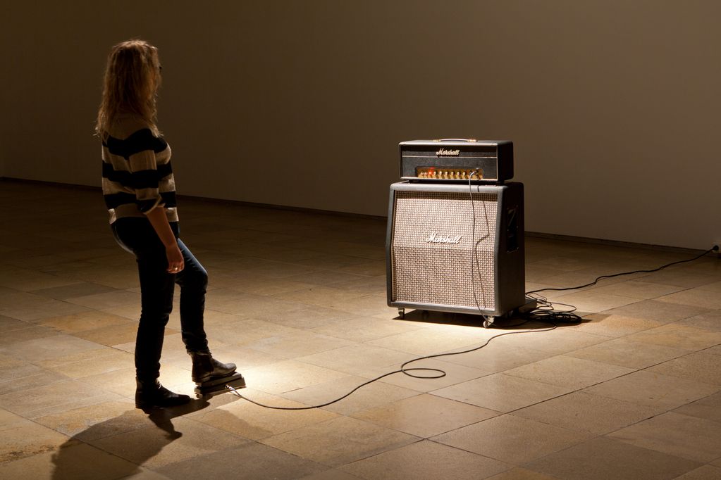 Diese Fotografie zeigt eine Frau in einem Raum mit einem Verstärker der Marke Marshall. Sie tritt auf ein Pedal das mit dem Verstärker verbunden ist und scheint ihn so betätigen zu können. Janet Cardiff/George Bures Miller, Sammlung Goetz München