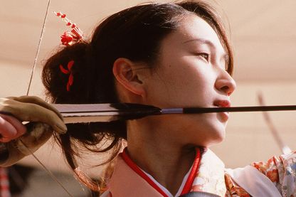 Nahaufnahme einer jungen asiatischen Frau, deren Gesicht im Profil von einer unteren Warte aus zu sehen ist. Ihre rechte Hand spannt gerade einen Pfeil in einem Bogen, sie selbst ist auf ihr Ziel fokussiert. Fiona Tan, Sammlung Goetz München