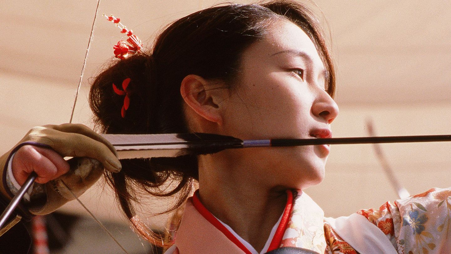 Close-up of a young Asian woman whose face is seen in profile from a lower vantage point. Her right hand is just stretching an arrow in a bow, she herself is focused on her target. Fiona Tan, Sammlung Goetz Munich