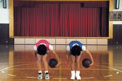 Dieses weitere Filmstill der Arbeit "Goshogaoka" von Sharon Lockhart zeigt die Frontalansicht zwei Basketballspieler(*innen), der eine in einem roten, der andere in einem blauen Trikot den gleichen Trick in gleicher Haltung ausführend in einer Schulturnhalle mit einer Theaterbühne samt zugezogenen Vorhang im Hintergrund.