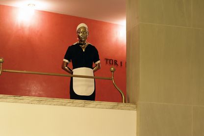 Ausstellungsansicht, die die Arbeit Tala in einem Opernhaus im Foyer zeigt. Die Arbeit besteht aus der Messingskulptur eines Hausmädchens in typischer Kluft (schwarzes Kleid, weiße Schürze und Haube). Elmgreen/Dragset, Sammlung Goetz München