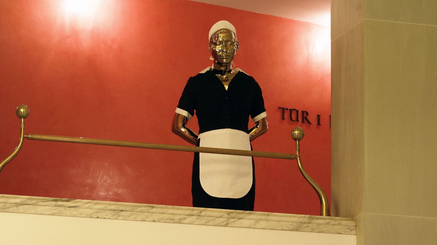 Ausstellungsansicht, die die Arbeit Tala in einem Opernhaus im Foyer zeigt. Die Arbeit besteht aus der Messingskulptur eines Hausmädchens in typischer Kluft (schwarzes Kleid, weiße Schürze und Haube). Elmgreen/Dragset, Sammlung Goetz München
