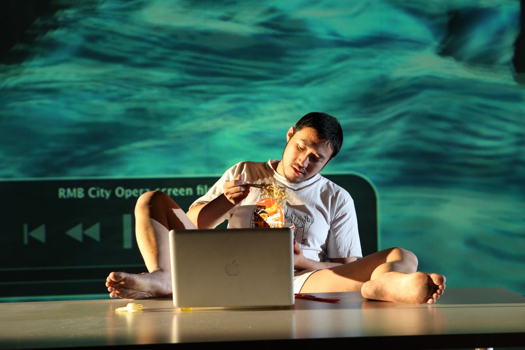Video Still eines jungen asiatischen Mannes, der vor einem silbernem Apple-Laptop sitzt, die Füße auf dem Tisch abgelegt hat und Instant Nudeln aus einem Plastikbehälter mit Stäbchen isst. Cao Fei, Sammlung Goetz München