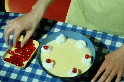 Dieses Video Still zeigt den Ausschnitt einer Draufsicht auf die Hände einer weiblichen Person, die einen Nachtisch in einer Schüssel mit roten Früchten garniert. Matthias Müller, Sammlung Goetz München