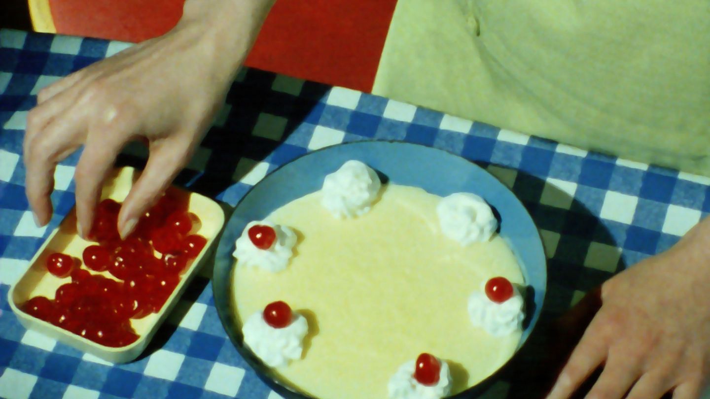 This video still shows a section of a top view of the hands of a female person garnishing a dessert in a bowl of red fruit. Matthias Müller, Sammlung Goetz Munich