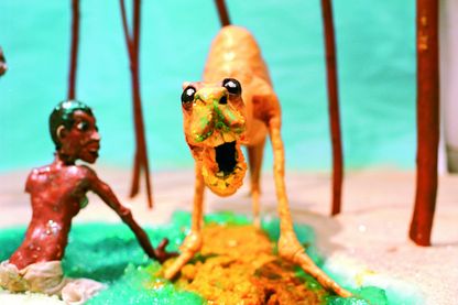 Dieses Video Still zeigt ein Kamel in einer Oase, neben ihm sitzt ein schwarzer Junge im türkisblauen Wasser. Es hat das Maul weit geöffnet und blickt mit einer paradoxen Mischung aus Freundlichkeit und Aggression in die Kamera. Beide Figuren sowie das Setting bestehen aus Knetmasse. Nathalie Djurberg & Hans Berg, Sammlung Goetz München