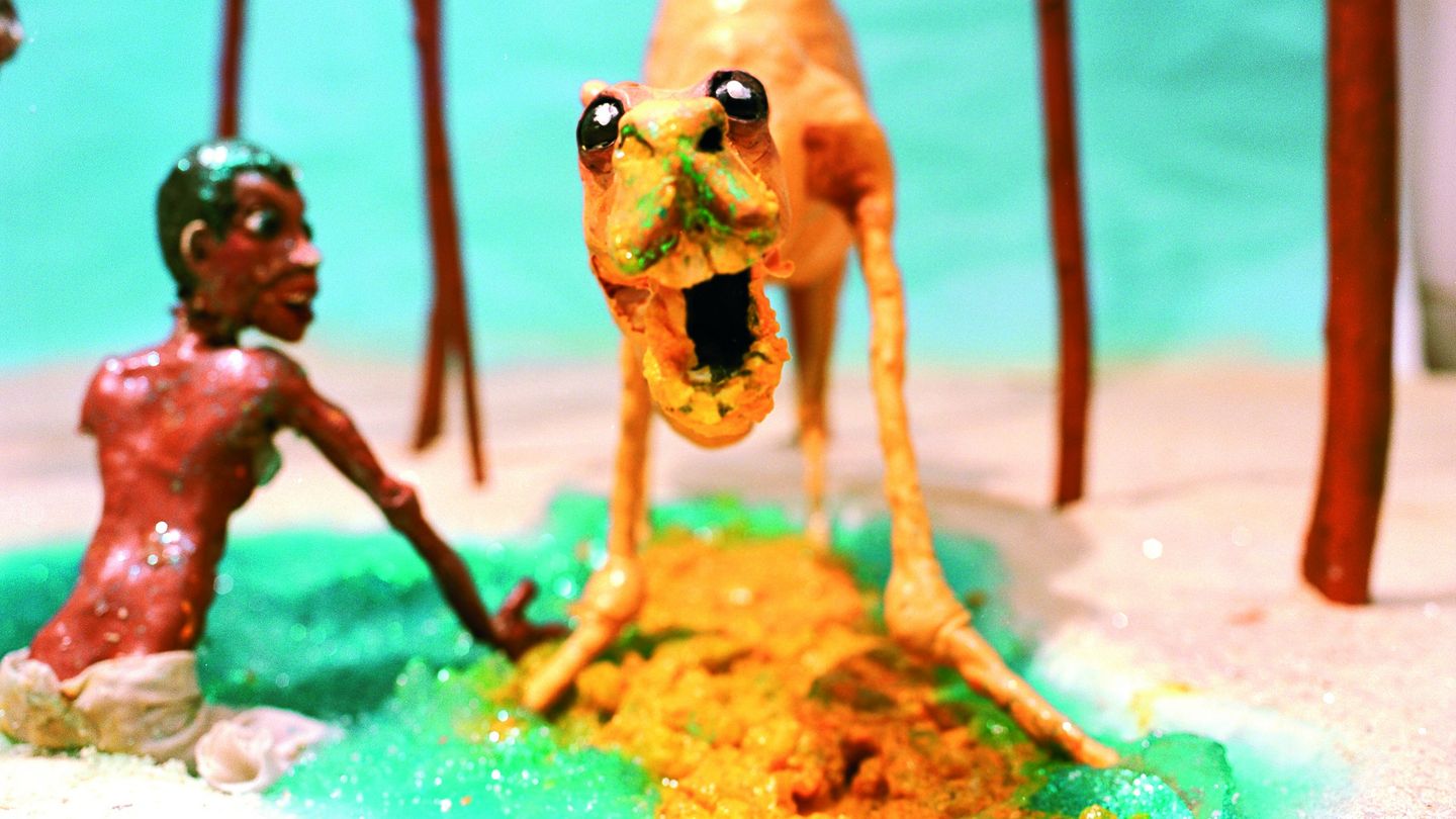 This video still shows a camel in an oasis, with a black boy sitting next to it in turquoise water. It has its mouth wide open and looks into the camera with a paradoxical mixture of friendliness and aggression. Both figures and the setting are made of plasticine. Nathalie Djurberg & Hans Berg, Sammlung Goetz Munich