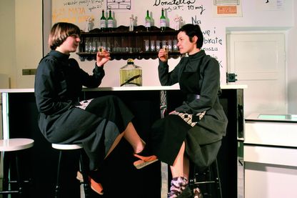 Diese Fotografie zeigt die Künstlerinnen Lucy McKenzie (links) und Paulina Olowska (rechts) an einer Bar sitzend mit gefüllten Schnapsgläsern, die sie zum Anstoßen erhoben haben. An der Wand hinter ihnen stehen Wörter, vermutlich Drinks, auf polnisch, dazwischen hängen einige Blätter aus der gemeinsamen Serie "Nova Popularna". Lucy McKenzie/Paulina Olowska, Sammlung Goetz München