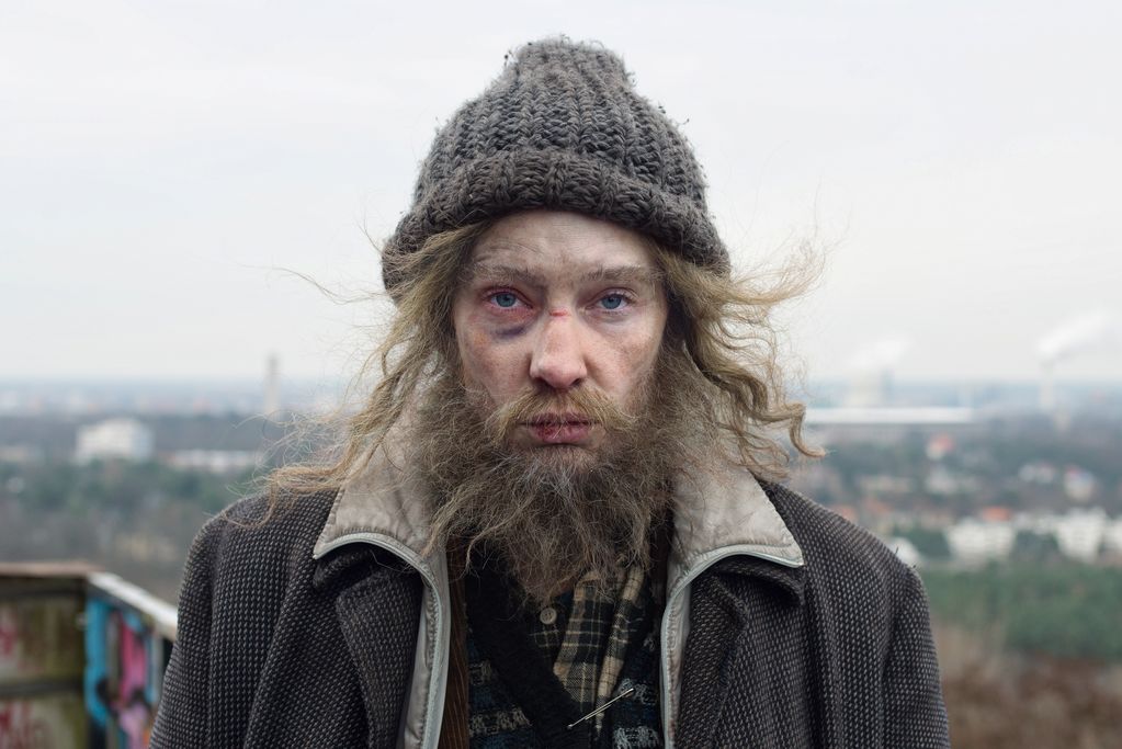 Man with dirty face and clothes as well as tangled hair peeking out from under a crocheted cap, looking into the camera with clear eyes. Julian Rosefeldt, Sammlung Goetz Munich