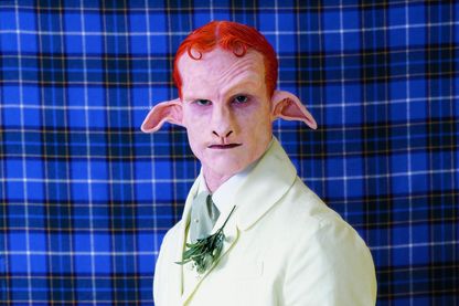 Auf diesem Production Still ist eine Art Satyr mit rotem Haar und weißem Anzug zu erkennen. Er steht vor einem Tartanstoff in blau.