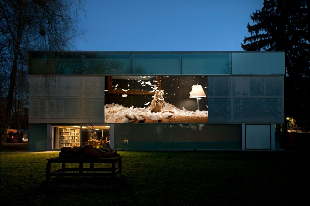 Nightly video projection onto the façade of the Sammlung Goetz. The video excerpt shows an owl in a furnished interior. On the right edge of the image, the light of a living room lamp is on. There are white feathers on the floor, which also fly around in the air.  