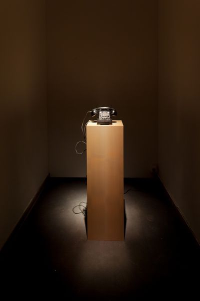Zu sehen ist ein altmodisches schwarzes Kabeltelefon mit Wählscheibe, auf einem quadratischen Holzsockel in einem dunklen Raum, von einem Spot beleuchtet. Janet Cardiff/George Bures Miller, Sammlung Goetz München