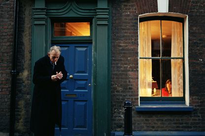 Video Still eines älteren Mannes in dunklem Mantel vor einer Haustür eines Backsteingebäudes, der im Begriff ist, sich eine Zigarette anzuzünden. Sam Taylor-Johnson, Sammlung Goetz München