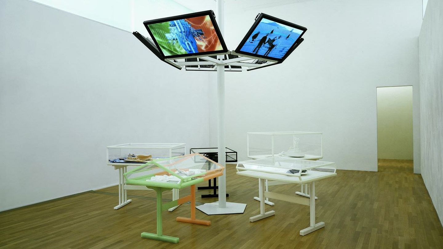 Fotografie der Installationsansicht des CREMASTER Zyklus in der Sammlung Goetz. Sechs Flachbildschirme sind an einem weißen Rohr in der Mitte des Raumes befestigt, darunter befinden sich Vitrinen in unterschiedlicher Ausführung mit verschieden Artefakten, die beispielsweise für die Ausstattung des Zyklus angefertigt wurden. Matthew Barney, Sammlung Goetz München