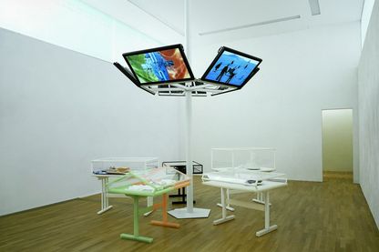 Fotografie der Installationsansicht des CREMASTER Zyklus in der Sammlung Goetz. Sechs Flachbildschirme sind an einem weißen Rohr in der Mitte des Raumes befestigt, darunter befinden sich Vitrinen in unterschiedlicher Ausführung mit verschieden Artefakten, die beispielsweise für die Ausstattung des Zyklus angefertigt wurden. Matthew Barney, Sammlung Goetz München