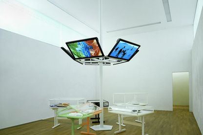 Photography of the installation view of the CREMASTER cycle in the Sammlung Goetz. Six flat screens are attached to a white duct in the center of the room, below which are vitrines of varying design containing various artifacts made, for example, to equip the cycle. Matthew Barney, Sammlung Goetz Munich