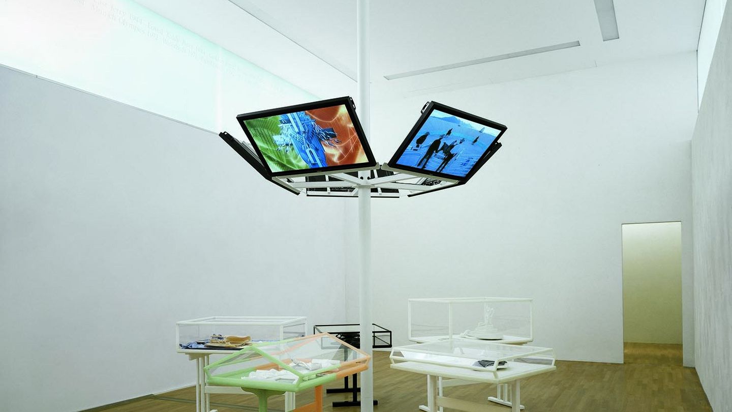 Photography of the installation view of the CREMASTER cycle in the Sammlung Goetz. Six flat screens are attached to a white duct in the center of the room, below which are vitrines of varying design containing various artifacts made, for example, to equip the cycle. Matthew Barney, Sammlung Goetz Munich