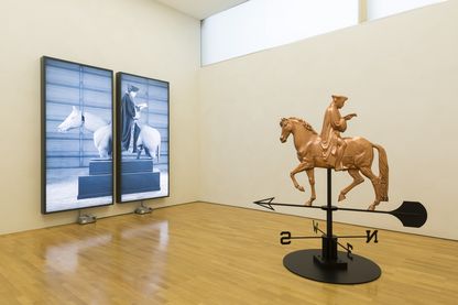 Ausstellungsansicht zweier Arbeiten des Künstlers Rodney Graham im Gebäude der Sammlung Goetz. Die eine Arbeit ist ein fotografisches Dyptichon in schwarzweiß eines Reiterstandbildes, welches als skulpturaler Dachschmuck mit Himmelsrichtungsanzeigern in der Mitte des Raumes steht. Rodney Graham, Sammlung Goetz München