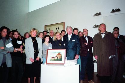 Die Fotografie zeigt die Besucher und Besucher*innen der Ausstellung in den Räumlichkeiten, dicht aneinander gedrängt. Im Vordergrund steht eine Fotografie auf einem weißen Sockel, hinter dem Publikum befindet sich eine Arbeit von Jannis Kounellis an der Wand.