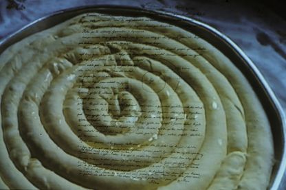 Diese Aufnahme ist ein Filmstill des Künstlers Anri Sala. Es zeigt ein albanisches Byrek in einer Backform, welches mit dem Bild eines handschriftlichem Briefes überlagert ist.