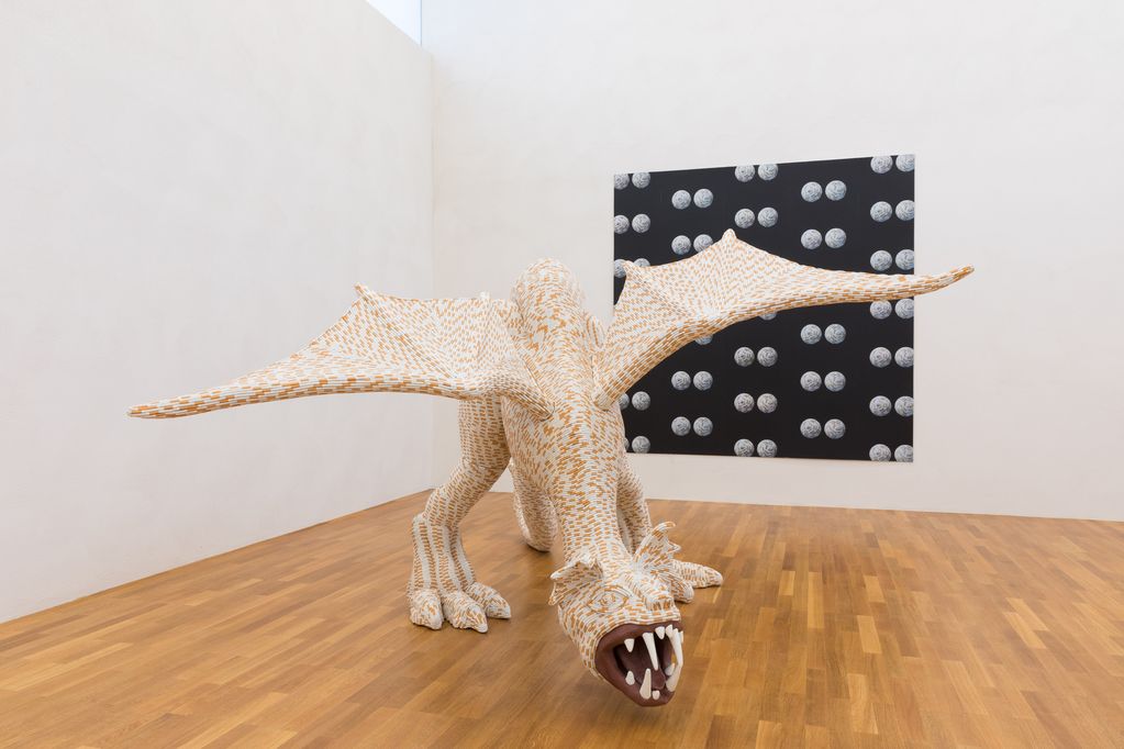 169 x 320 x 285 cm large dragon sculpture almost completely covered with cigarettes, on the wall behind it hangs a screen print with round objects also covered with cigarettes on a black background.  Sarah Lucas, Sammlung Goetz, Munich