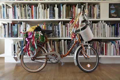 Kupferfarbenes Klapprad, behängt mit bunten Stoffbändern, einem Kanister, und einem Spielzeugschläger. Auf der Rückablage ist eine grüne Plastikkiste befestigt. Das Fahrrad steht vor einem Bücherregal, das ohne jegliche Ordnung einsortiert worden zu sein scheint. Andreas Slominski, Sammlung Goetz München