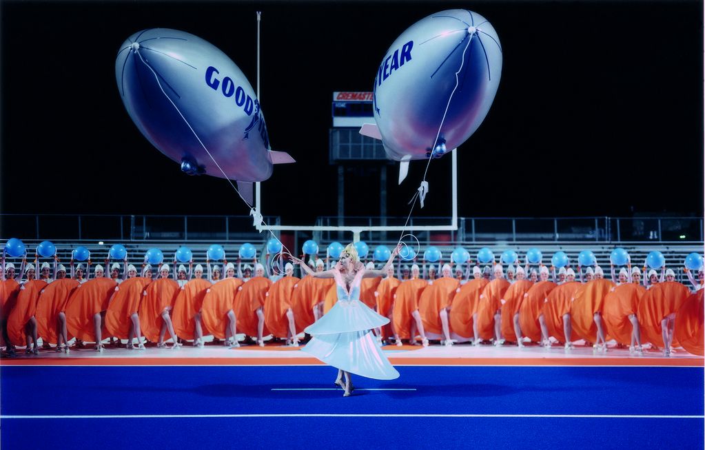 This film still shows a scene from the Cremaster cycle by American artist Matthew Barney. A woman in a futuristic dress holds two zeppelin balloons in her hands, with the words "Goodyear" written on them. In the background is a carefully lined up group of women holding their orange hoop skirts up in the same direction. The scene takes place on a track with a blue floor.