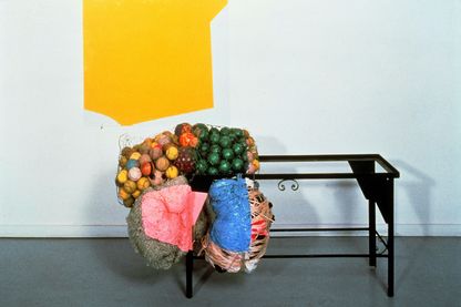 Diese Installationsaufnahme beinhaltet ein zwei- bis dreidimensionales Werk von Jessica Stockholder. Es besteht aus einer Acrylmalerei in Gelb auf Glas, einem schwarzen Eisengestell eines kleinen Tisches und bunten Bällen in Hasendraht, sowie eingewickelte Schafwolle und andere Textilien, die an dem Tischgestell angebunden sind. 