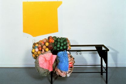 This installation shot contains a two to three dimensional work by Jessica Stockholder. It consists of an acrylic painting in yellow on glass, a black iron frame of a small table and colorful balls in rabbit wire, as well as wrapped sheep wool and other wrapped textiles tied to the table frame. 
