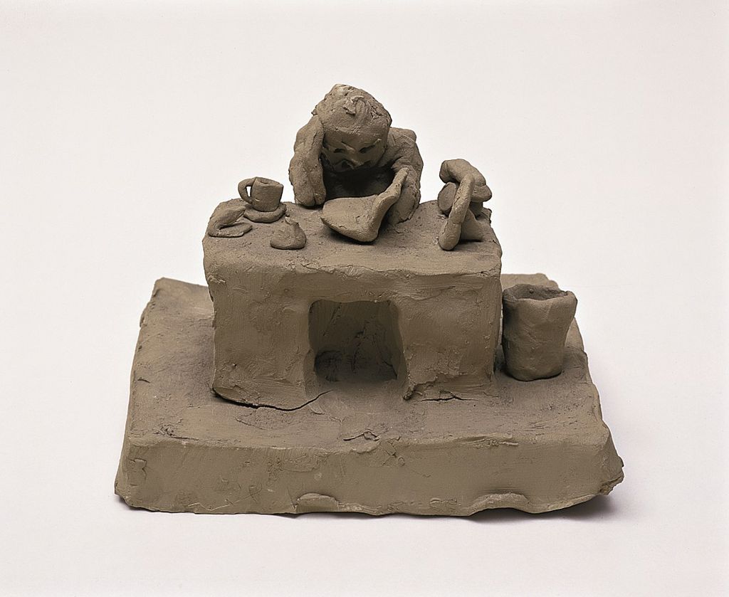 This photograph shows a childlike-looking clay sculpture of an accountant sitting at his desk surrounded by a coffee cup, punch, stapler, telephone and family photo.