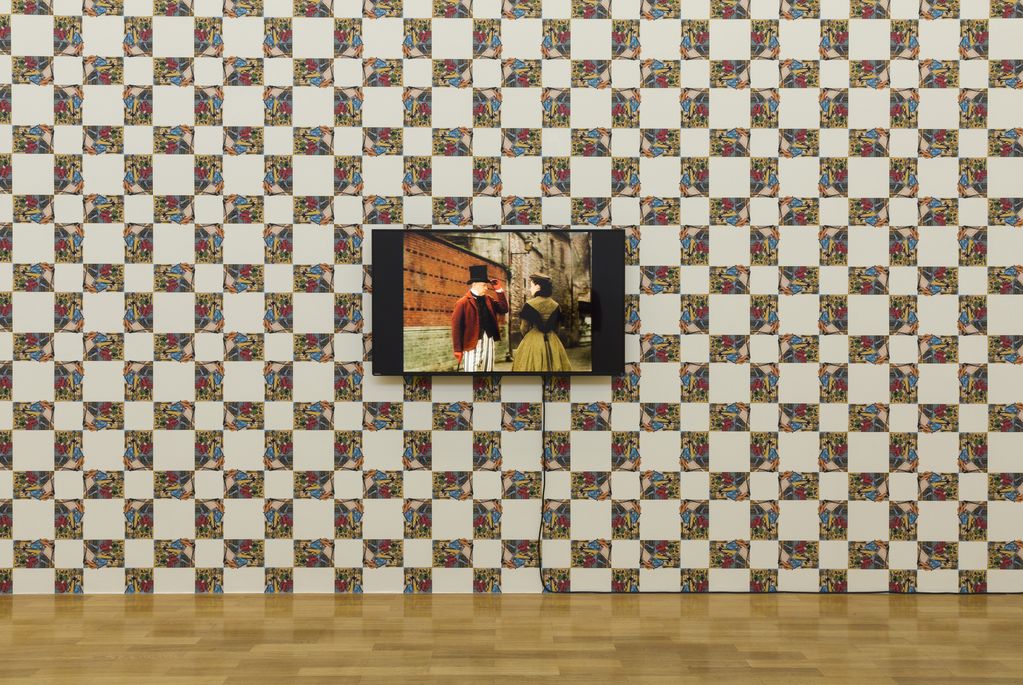 Ausstellungswand als Tapete mit wiederkehrendem Muster, auf der ein Bildschirm hängt, dessen Bildschirm einen Mann und eine Frau in historischer Kleidung zeigt. Rodney Graham, Sammlung Goetz München
