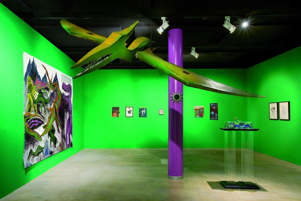Exhibition view showing a room arranged in green and purple. The column in the center is purple, the recessed walls are poisonous green, as is the flying dinosaur dangling from the ceiling. On the walls hang variously sized and colorful works by the artist. Andy Hope 1930, Sammlung Goetz Munich
