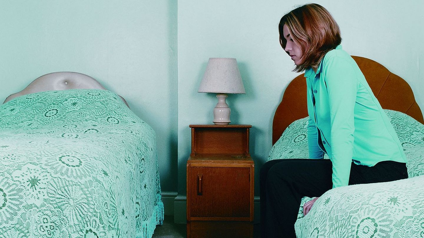 This colour photograph shows a woman in an old-fashioned looking room with two single beds. The young woman sits on one of the two beds and looks lost in thought at the other bed. The colourfulness of the photograph appears artificial and cold.