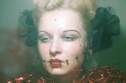 Portrait of a young, blond woman with 1920s make-up, a black net in front of her face and a ruff of red tinsel. Ulrike Ottinger, Sammlung Goetz Munich