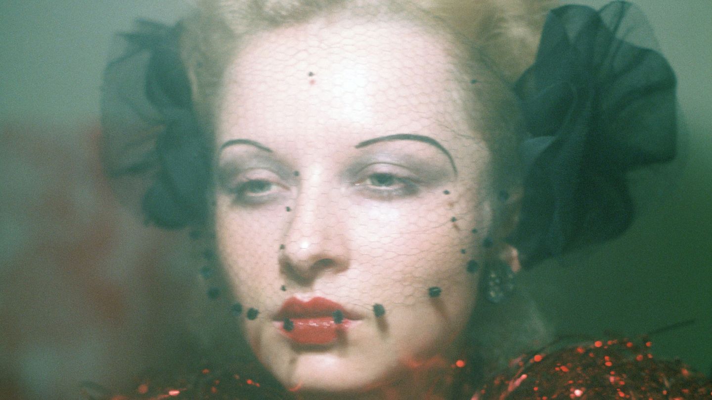 Portrait of a young, blond woman with 1920s make-up, a black net in front of her face and a ruff of red tinsel. Ulrike Ottinger, Sammlung Goetz Munich