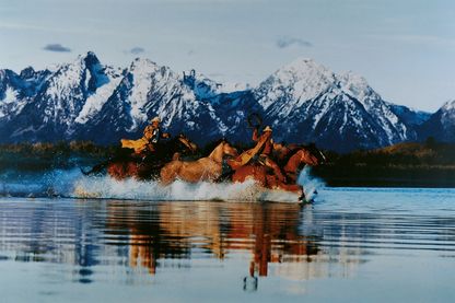 Diese Farbfotografie zeigt zwei Cowboys, die mit drei Pferden durch ein Gewässer vor verschneiter Gebirgslandschaft reiten. Richard Prince, Sammlung Goetz München